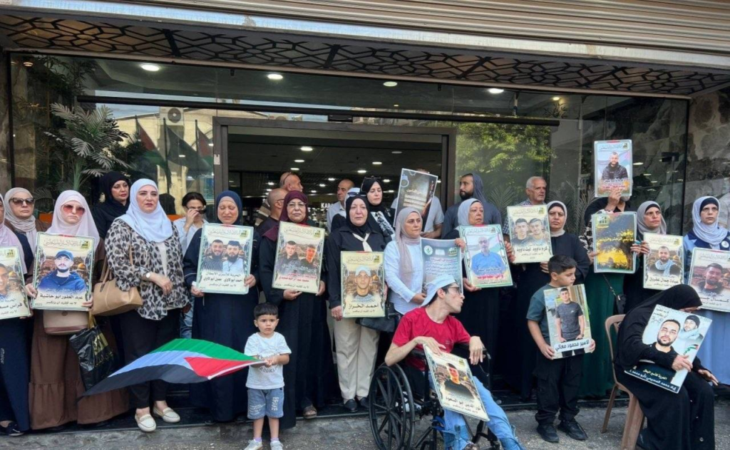 Families, most who are standing including women wearing hijabs, as well as some men, a few children, as well as a person in a wheelchair and a women sitting in a chair holding signs with photos of their family members detained by Israel.